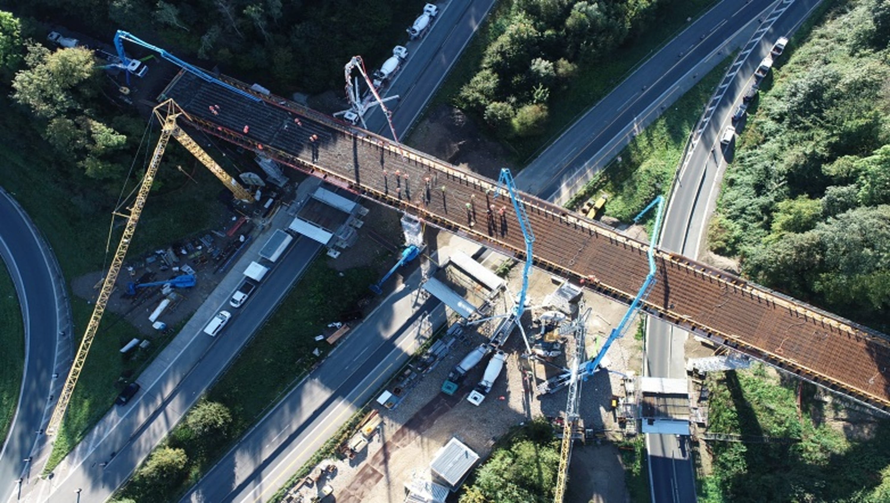 Die Luftaufnahme zeigt eine Brückenbaustelle mit mehreren Betonpumpen und Arbeitern in Warnwesten bei der Bewehrung. Umgeben ist das Geschehen von grüner Landschaft, Straße und Baubüros.
